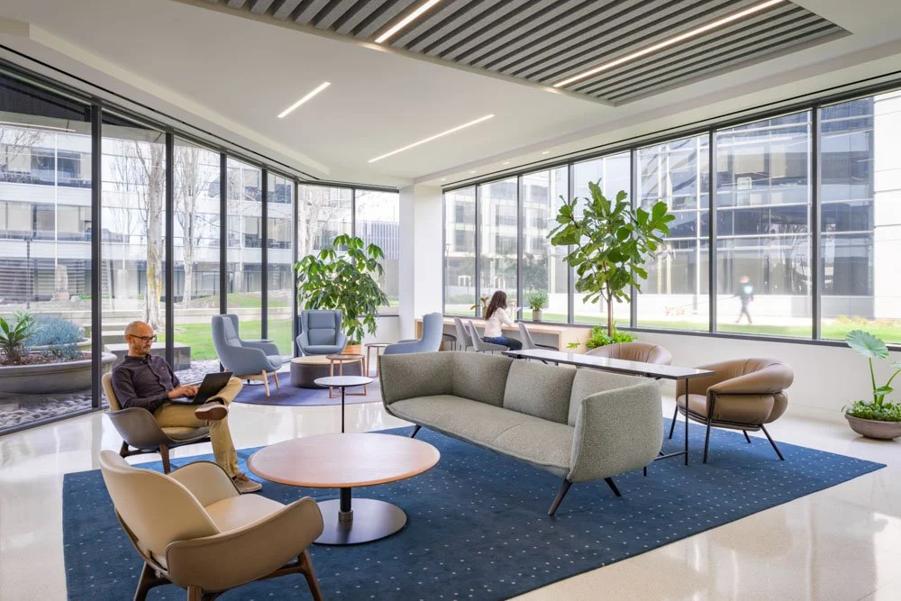 office lobby lounge area with modern sofas, chairs, and tables, large windows with ground level views, three people