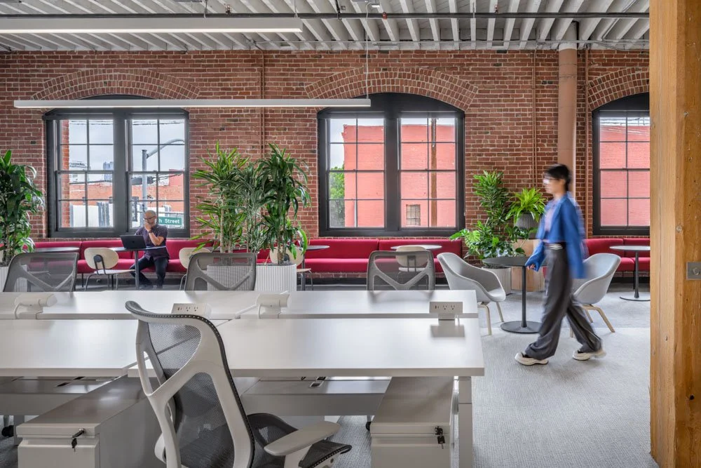 modern office with desks in foreground, brick wall and windows in background, two workers