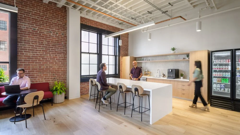 modern office kitchen with white island and bar seating, brick wall, light wood floor and cabinets, four people
