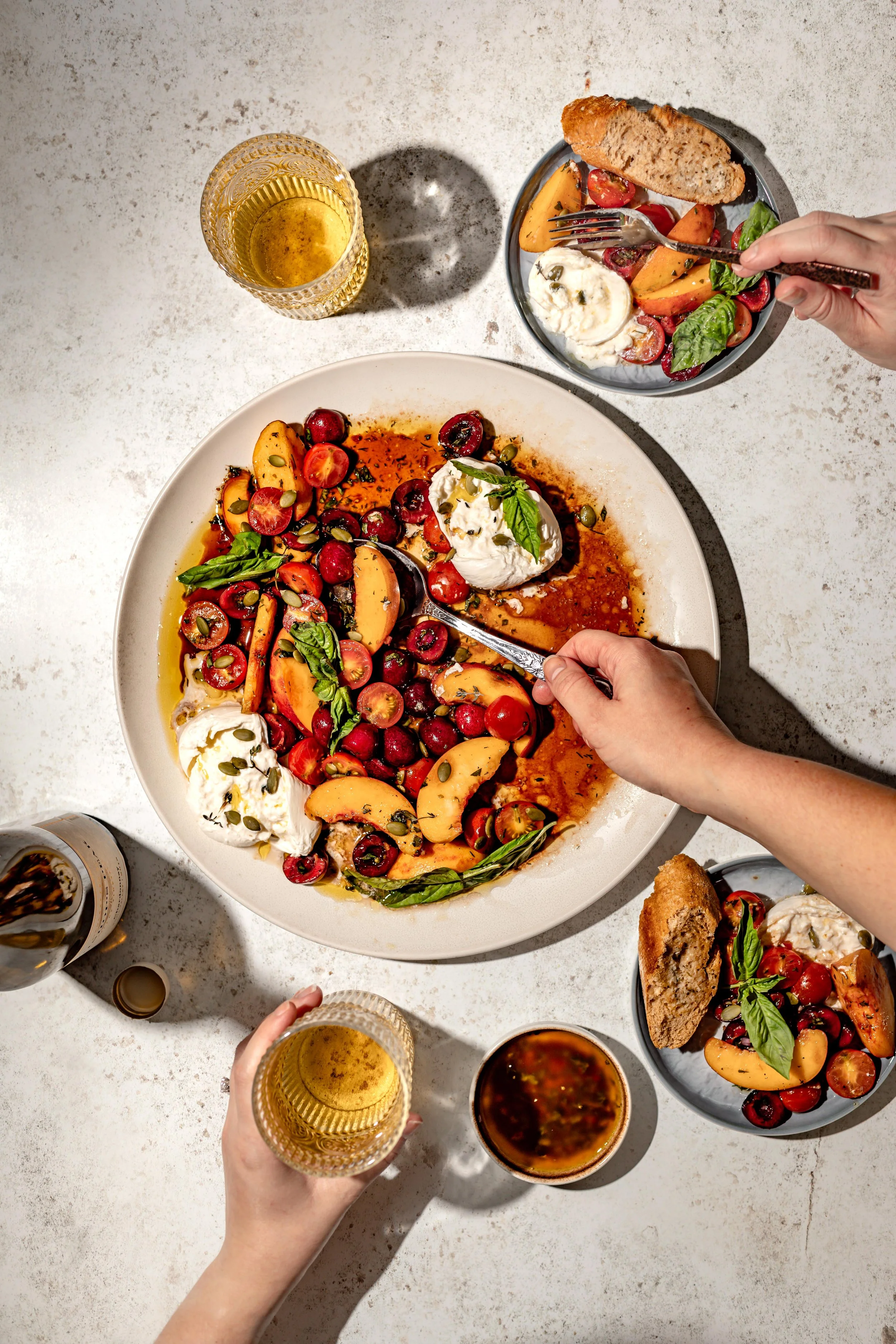 A top-down view of a shared meal with a large fruit and cheese salad, glasses of white wine, a small bowl of a dark sauce, and additional smaller bowls with fruit and bread, set on a white table.