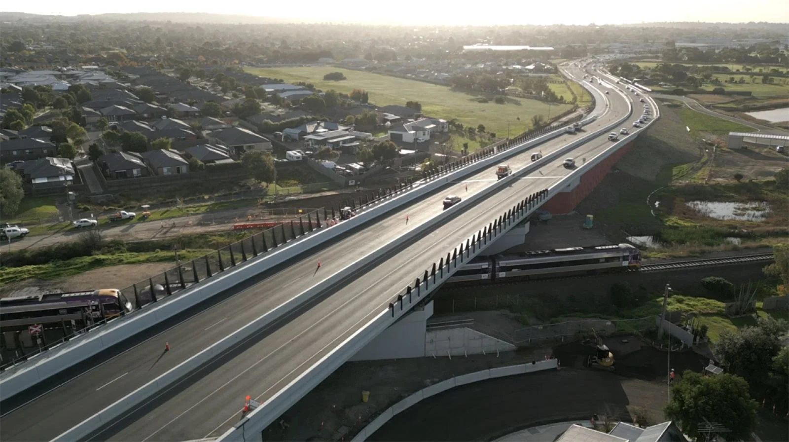 Aerial view of a new road bridge in Barwon Heads carrying traffic over a railway corridor in a suburban area, with trains passing below and open land to the side.