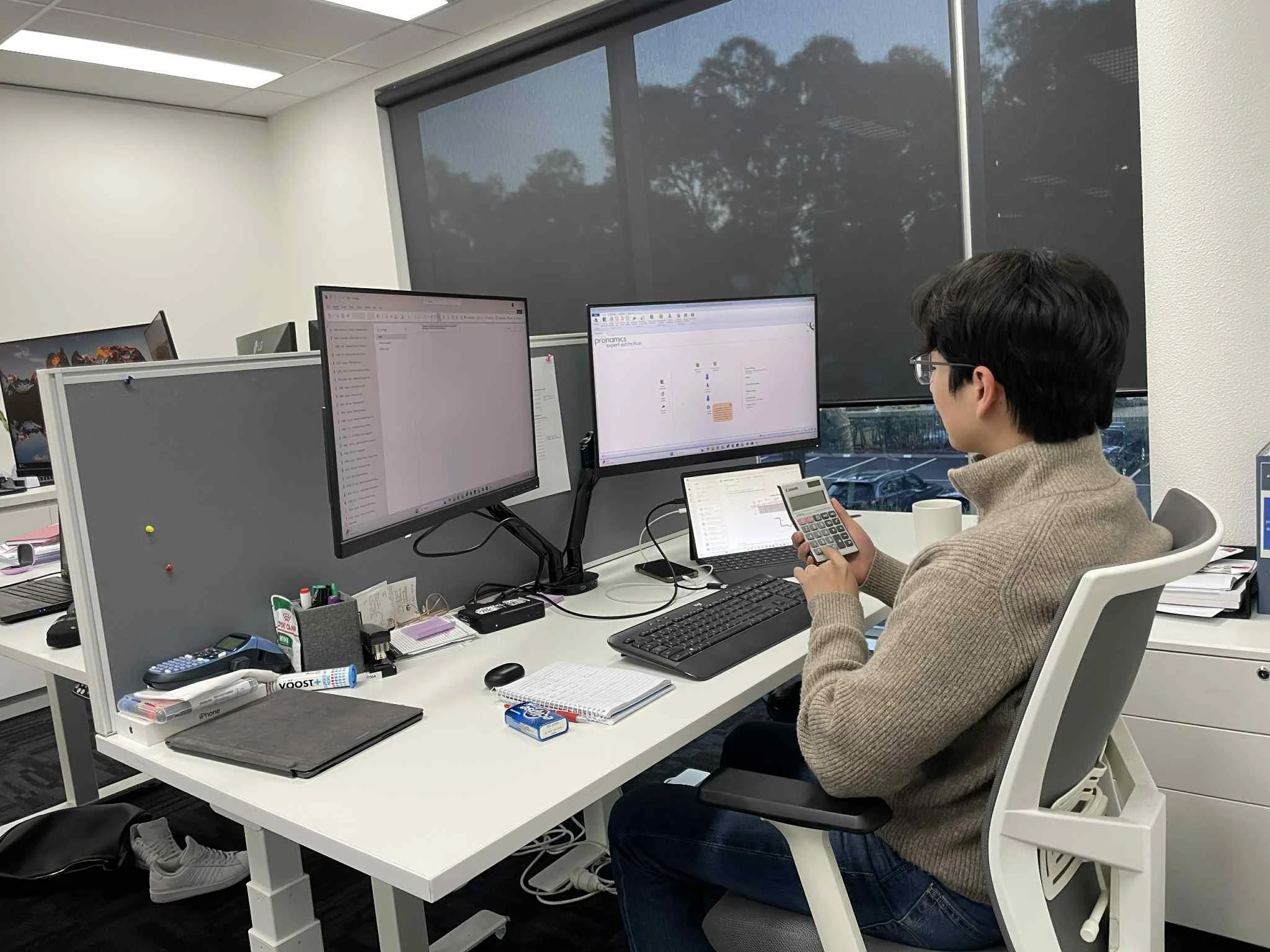 A man in a tan sweater sitting at a white office desk with two large monitors, a laptop, a calculator, and office supplies, working in an office with large windows and closed blinds.