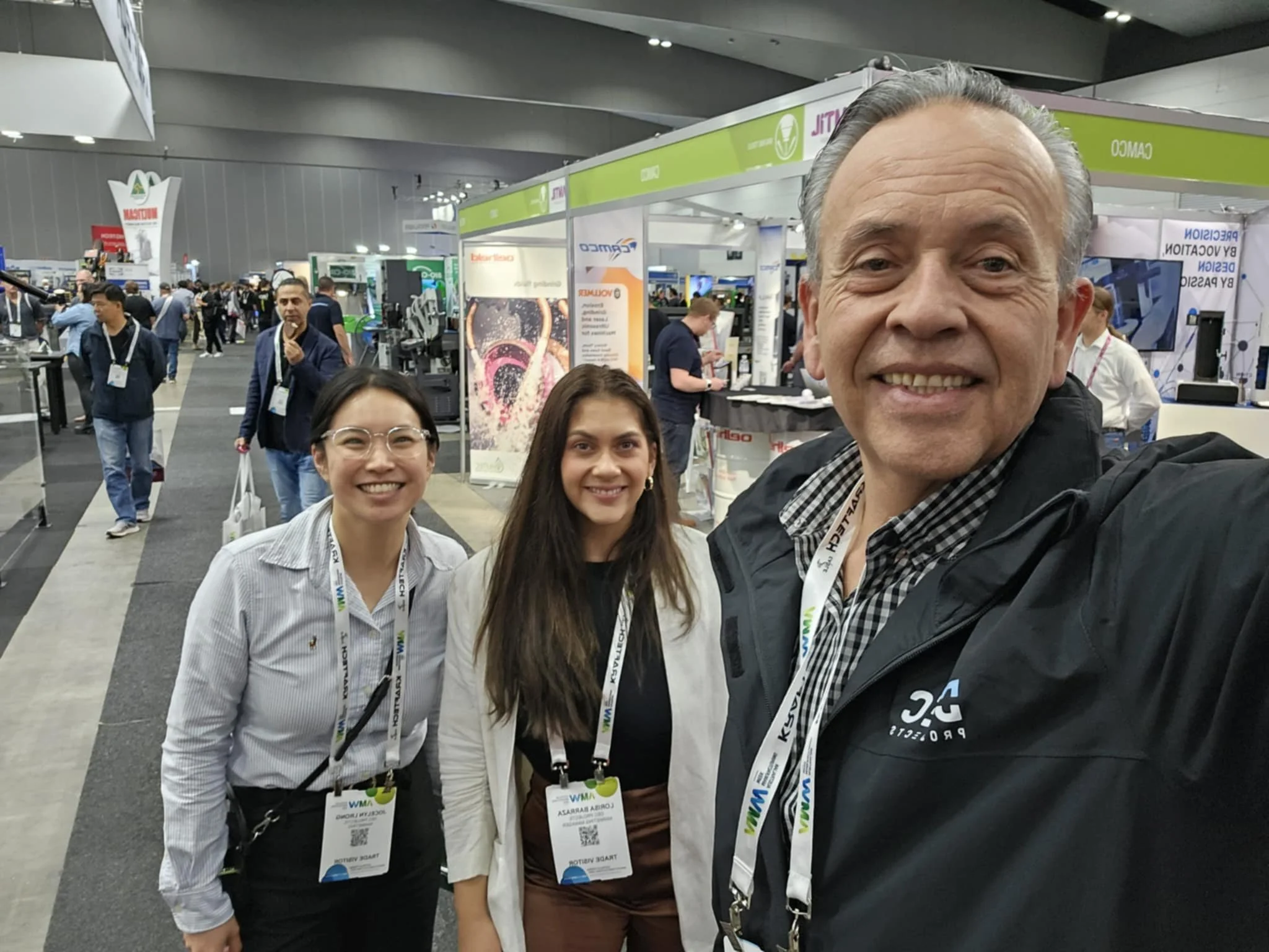 Three smiling people at a trade show or conference in Melbourne, with booths and other attendees in the background.