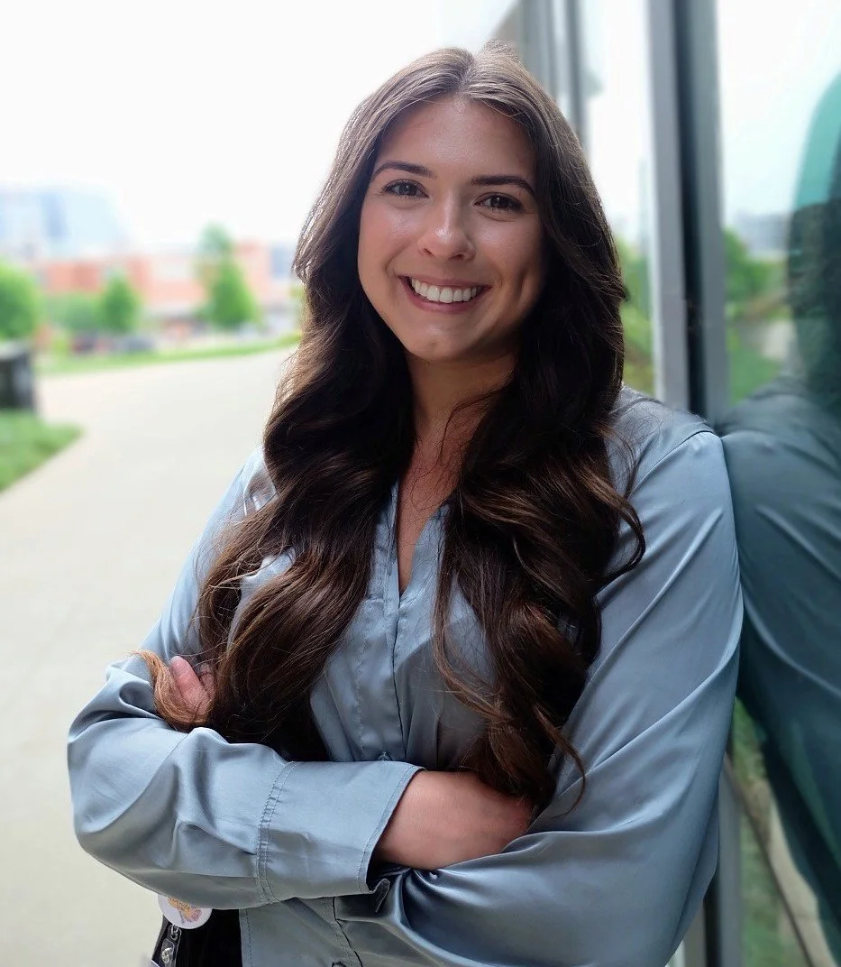 A young woman with long, wavy brown hair, smiling, wearing a light gray jacket, standing outdoors near a glass window with a blurred background of trees, buildings, and a walkway.