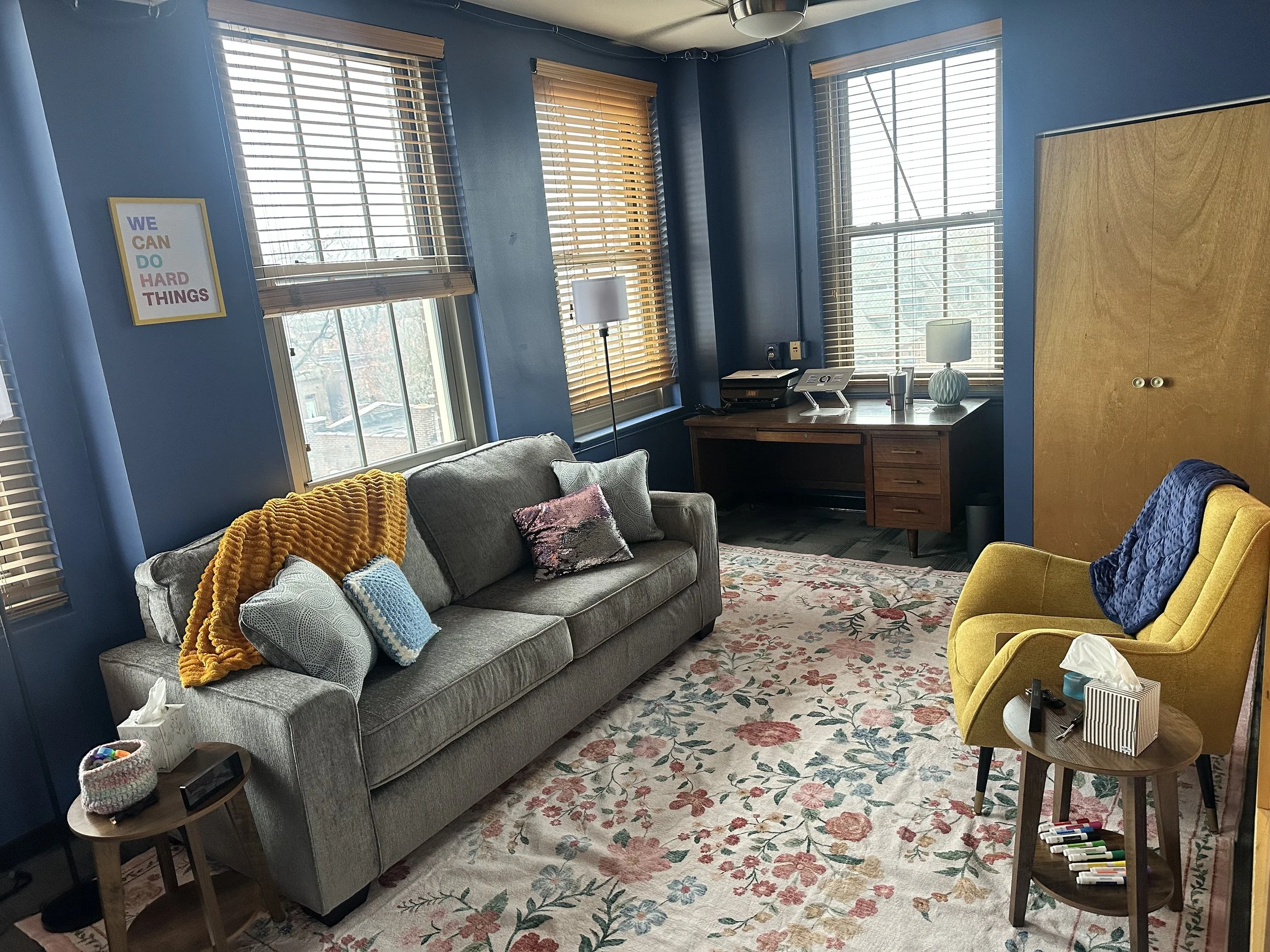 A cozy living room with blue walls, three windows with wooden blinds, a gray sofa with decorative pillows and a yellow throw blanket, a yellow armchair with a blue quilt, a wooden side table with tissue box and markers, a wooden desk with a lamp and office supplies, a framed motivational quote on the wall saying 'WE CAN DO HARD THINGS,' and a floral carpet.