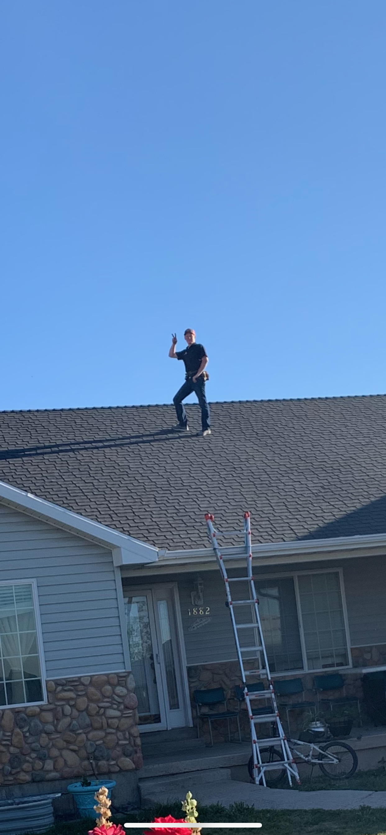 Person standing on the roof of a house making a peace sign with one hand, with a ladder leaning against the house, in front of a clear blue sky.