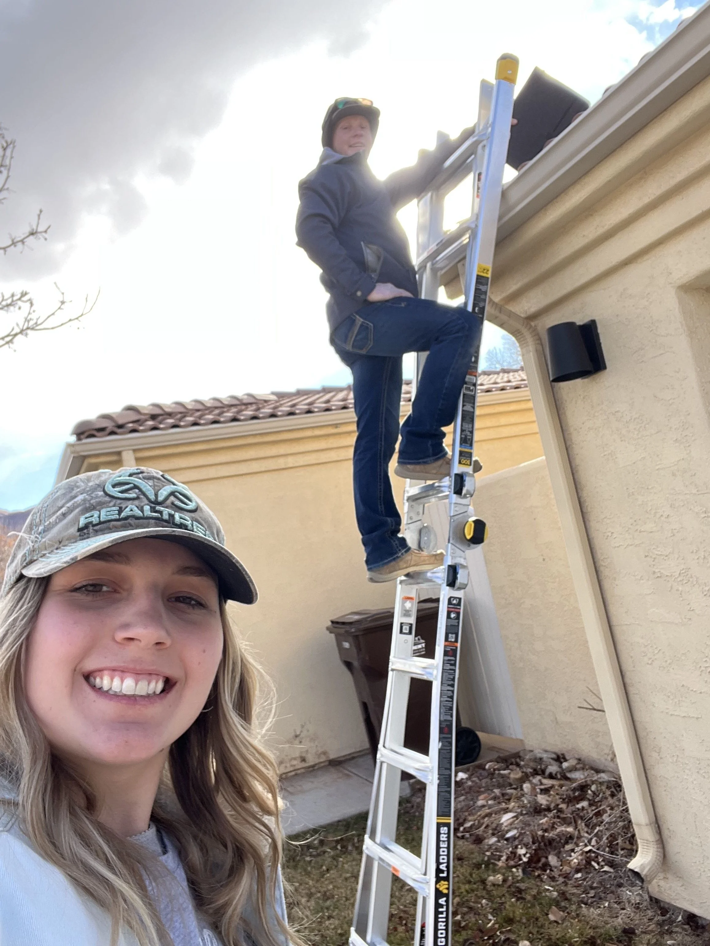 Two women engaged in roofing work; one woman takes a selfie smiling in the foreground, while the other woman stands on a ladder working near the roof of a house.