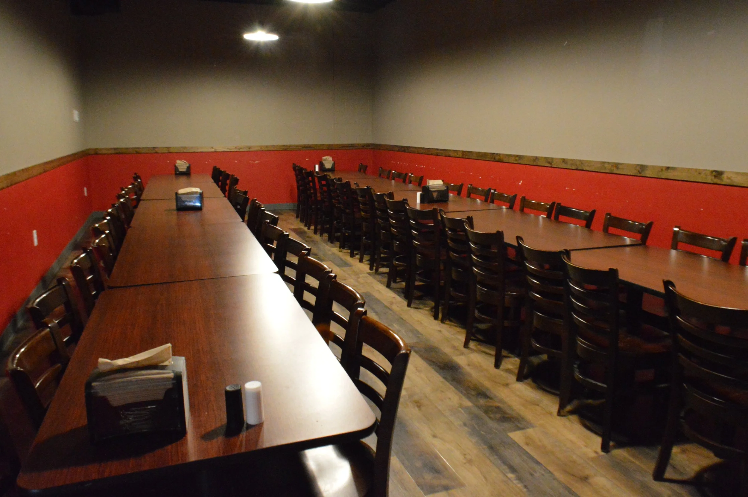 Empty restaurant or banquet hall with long wooden tables and chairs, napkin dispensers, salt and pepper shakers, with dim lighting and a red and beige wall.