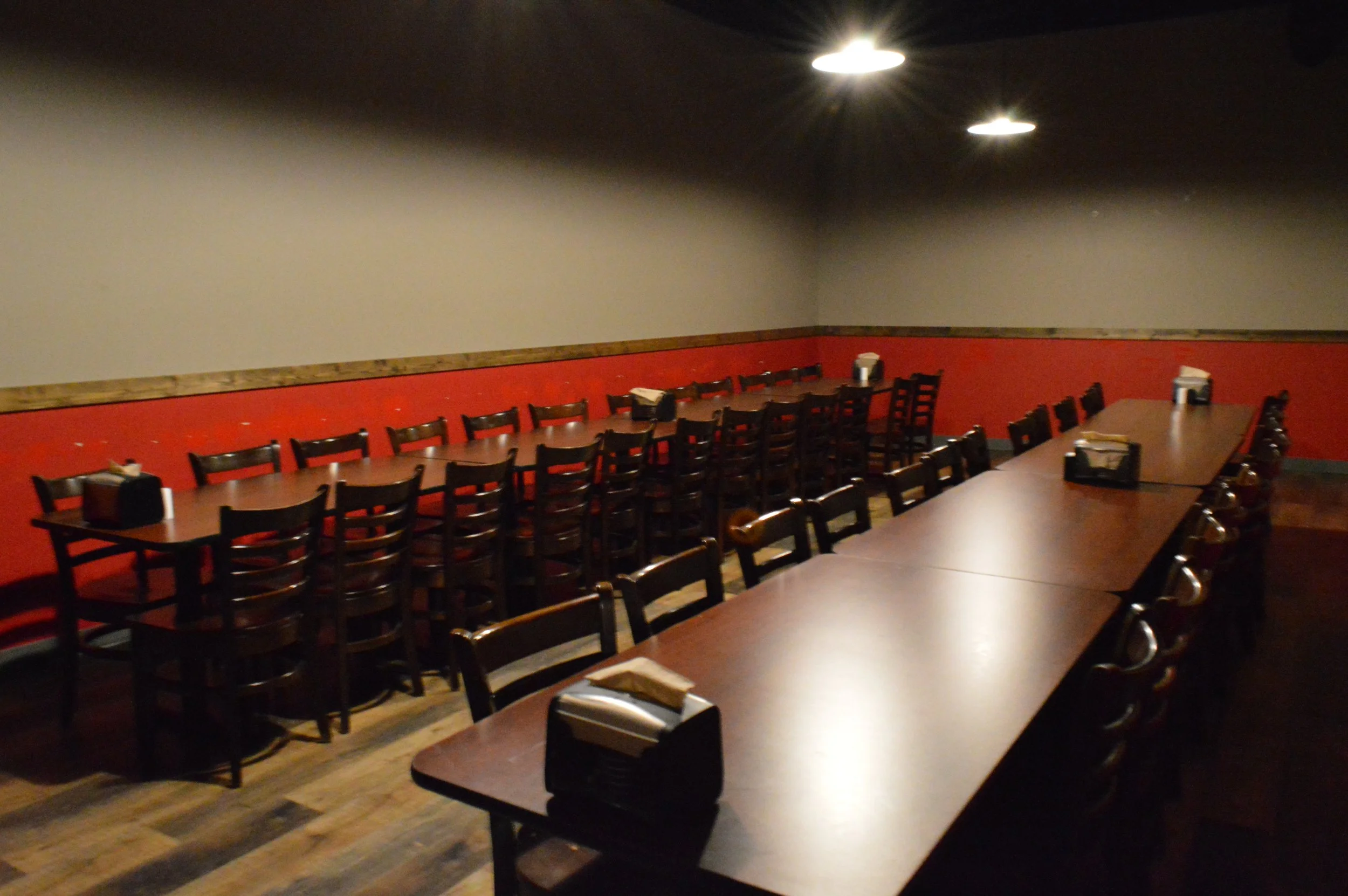 Empty restaurant with long tables, chairs, and napkin dispensers, dim lighting, and a red and gray wall.