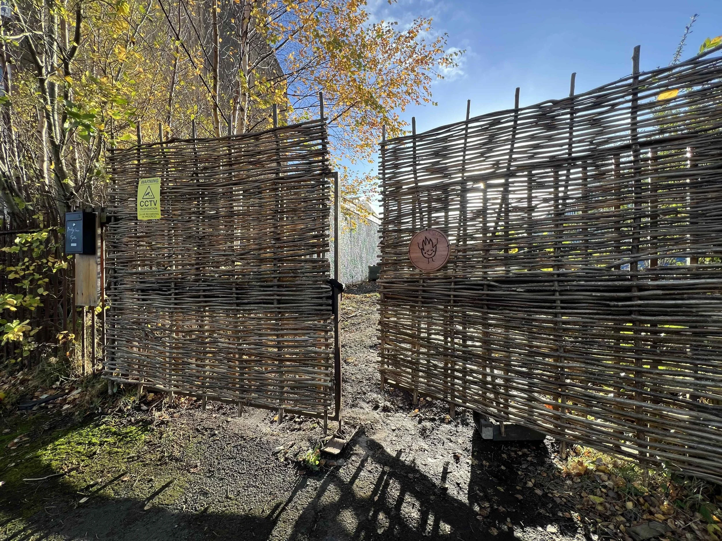 The front gate of Glasgow Community Sauna. It is a wooden gate with the Glasgow Community Sauna logo etched into a separate piece of wood. The entry gate is on McCulloch Street, Pollockshields