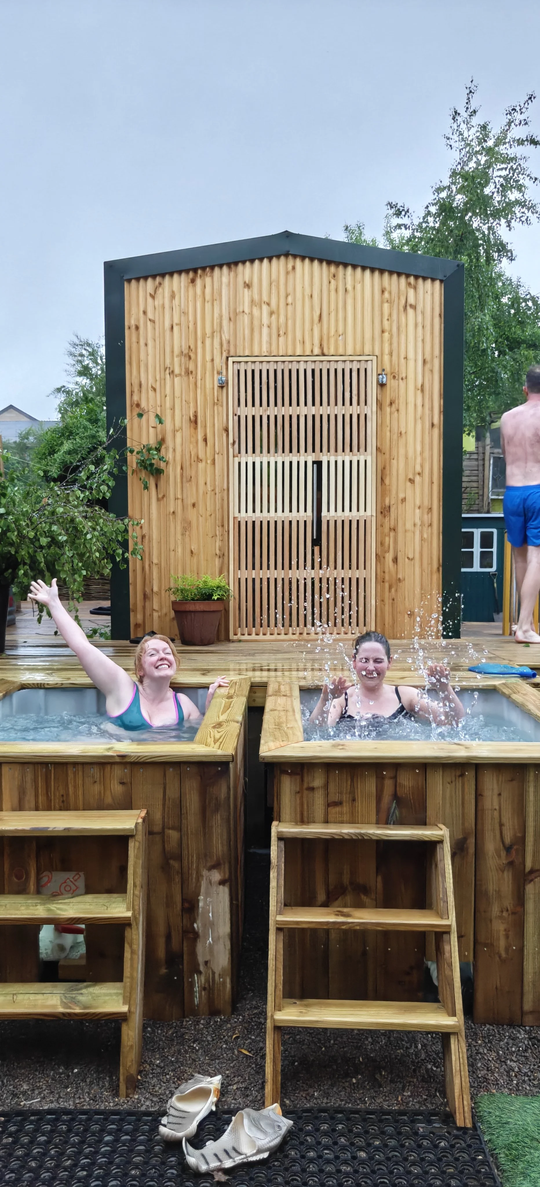 Two women enjoying hot tub outdoors with wooden structure and shower in the background, and a shirtless man walking away.