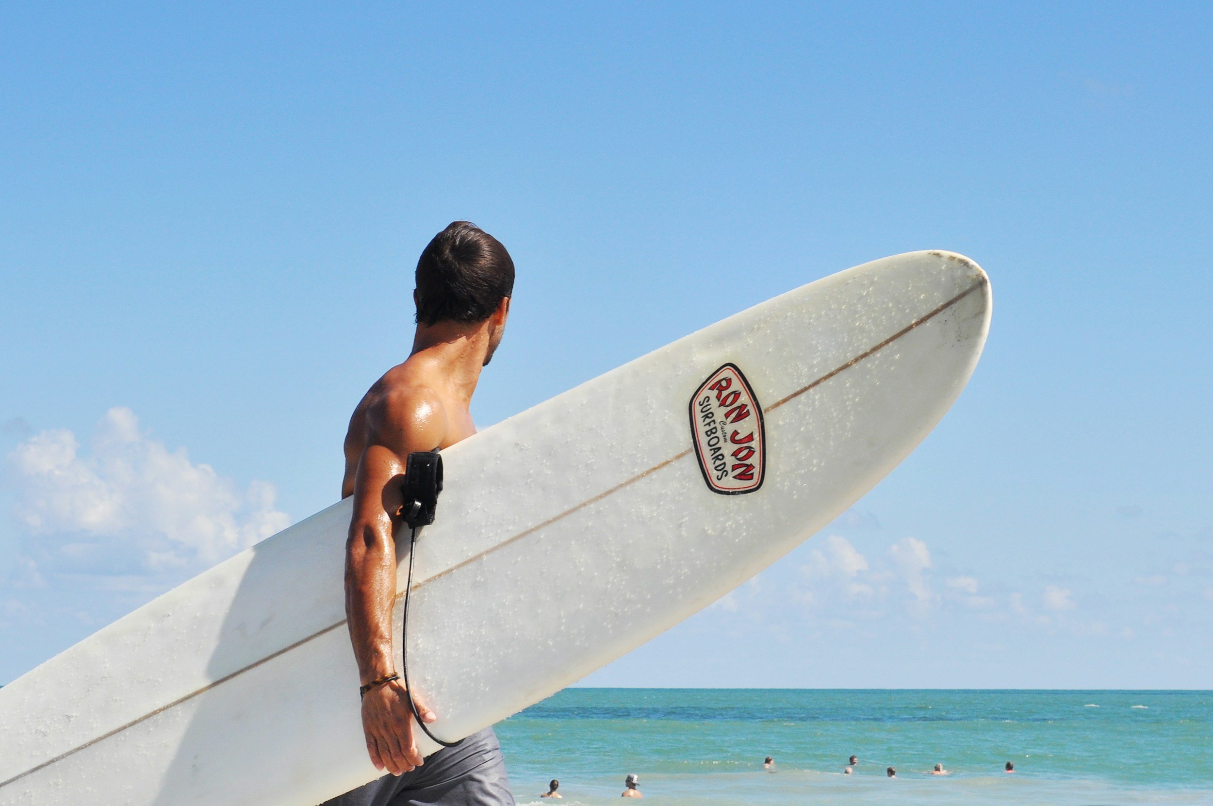 A man with tan skin and dark hair carries a white surfboard with a red and black logo while walking on the beach, with the ocean and blue sky in the background.