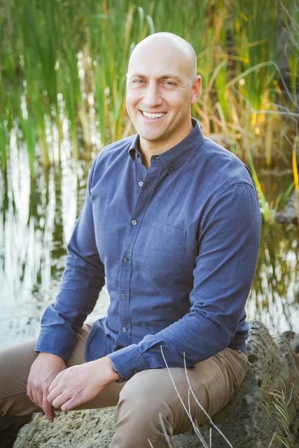 A bald man in a blue button-up shirt and beige pants sitting on a rock near a body of water, smiling with tall green grass in the background.