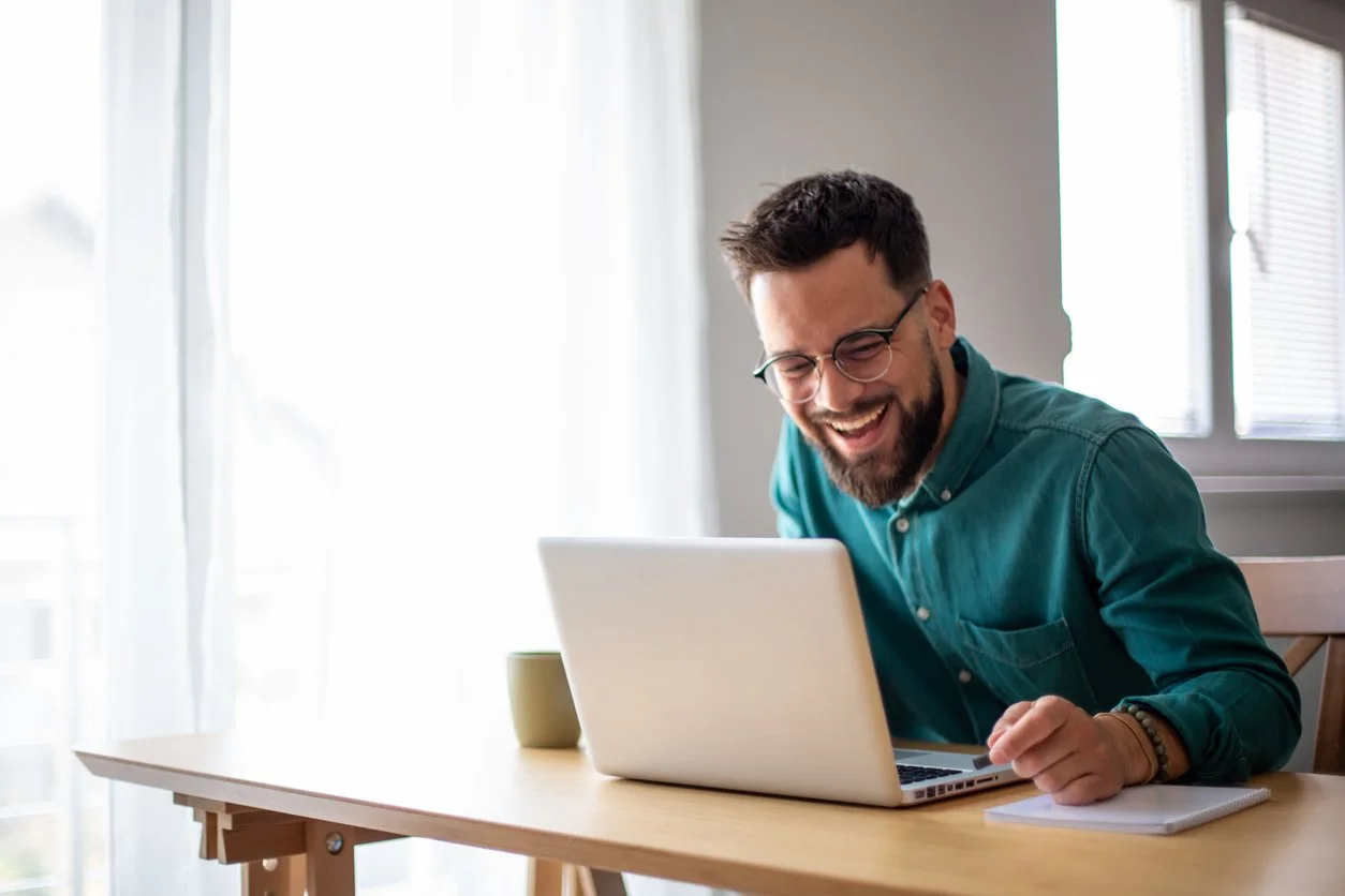 Man with glasses and a beard laughing while working on a laptop at a wooden table in a bright room.
