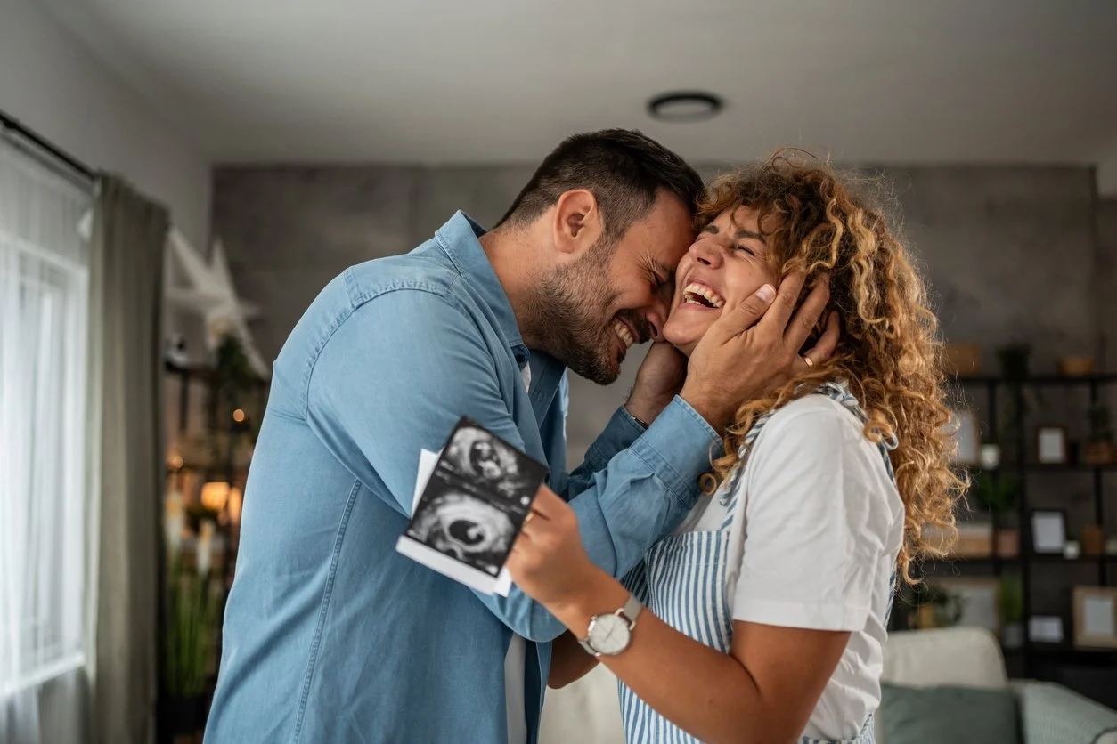 A couple celebrating pregnancy, with the woman holding an ultrasound image and the man touching her face, both smiling and joyful.