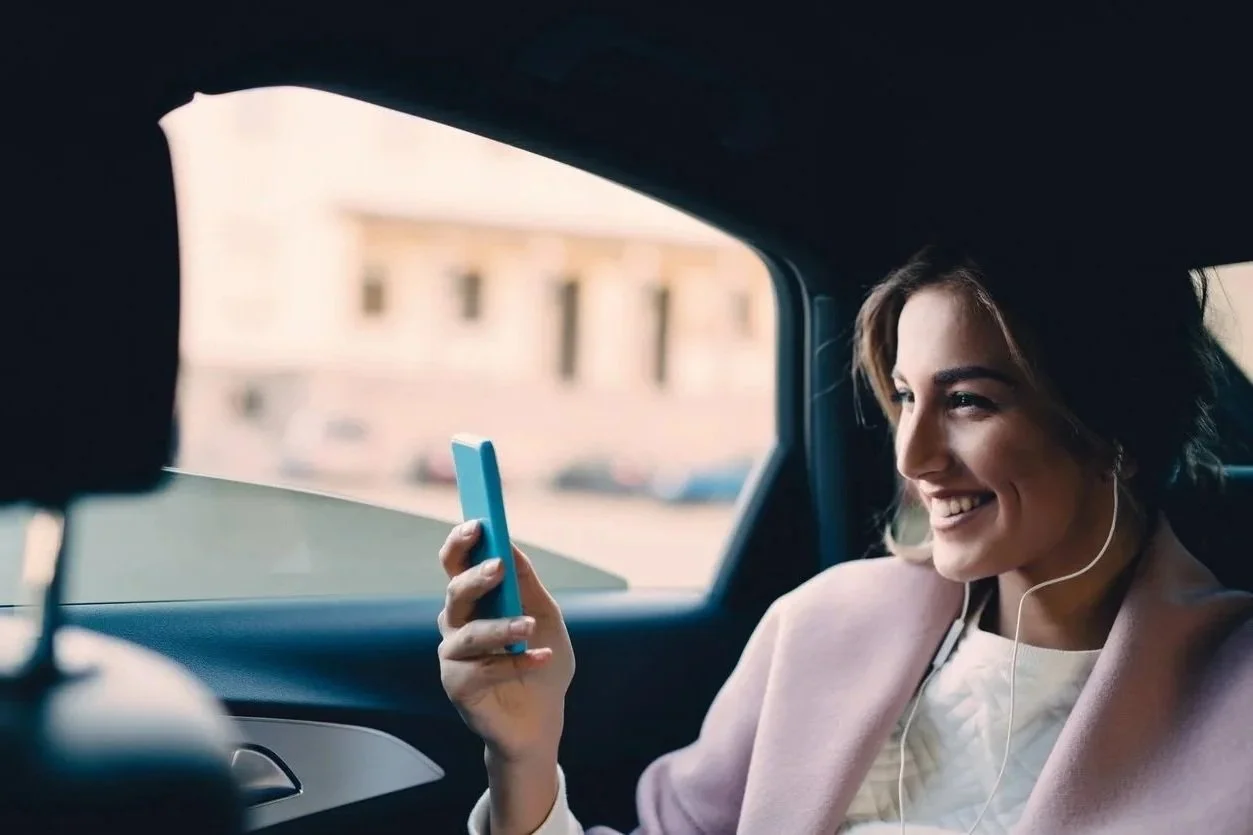 A woman sitting in the backseat of a car, smiling and holding a smartphone, with earphones in her ears.