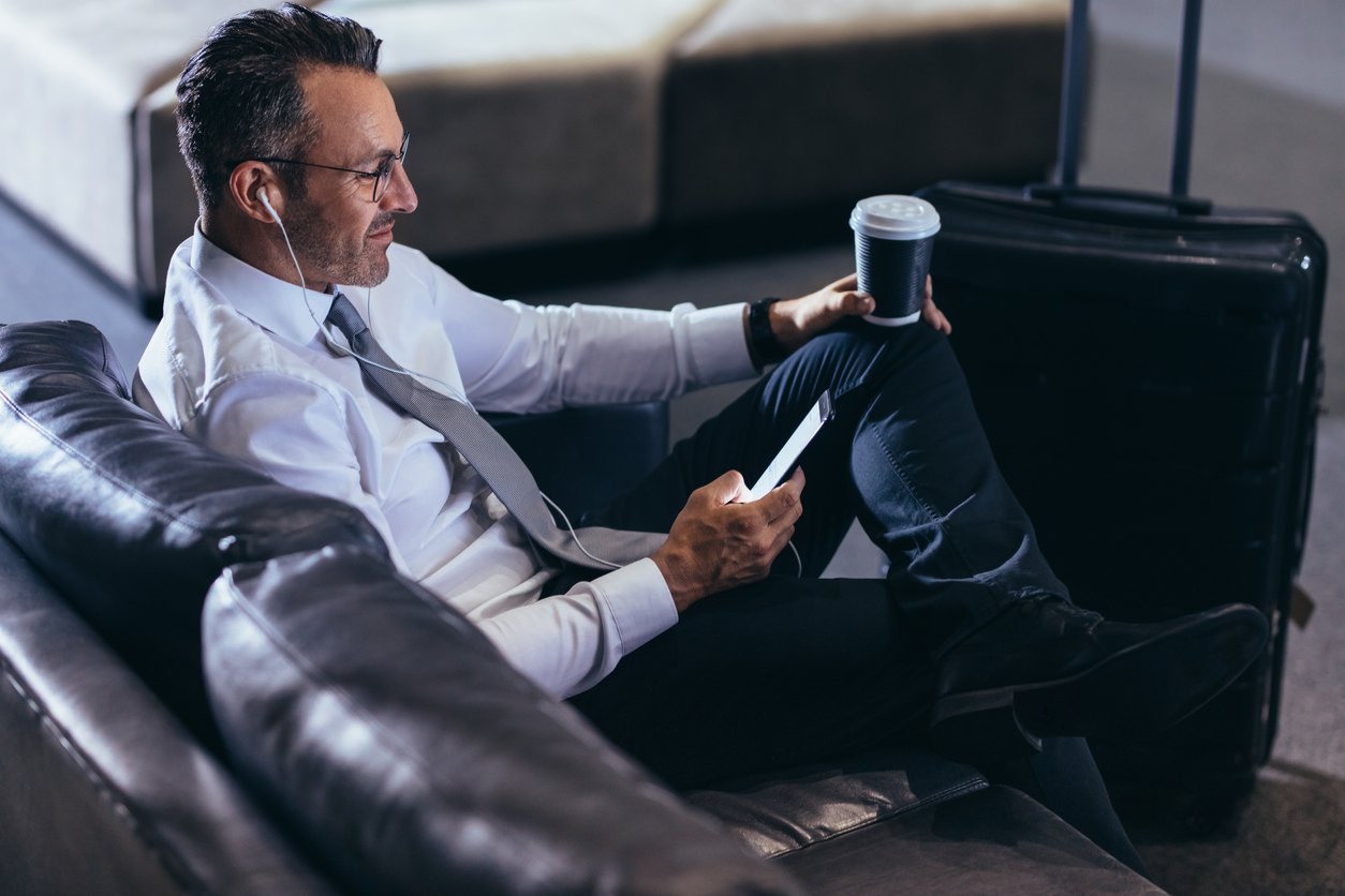 A man in business attire sitting on a leather couch at an airport, looking at his phone, with a suitcase and a coffee cup nearby, wearing earphones and smiling.