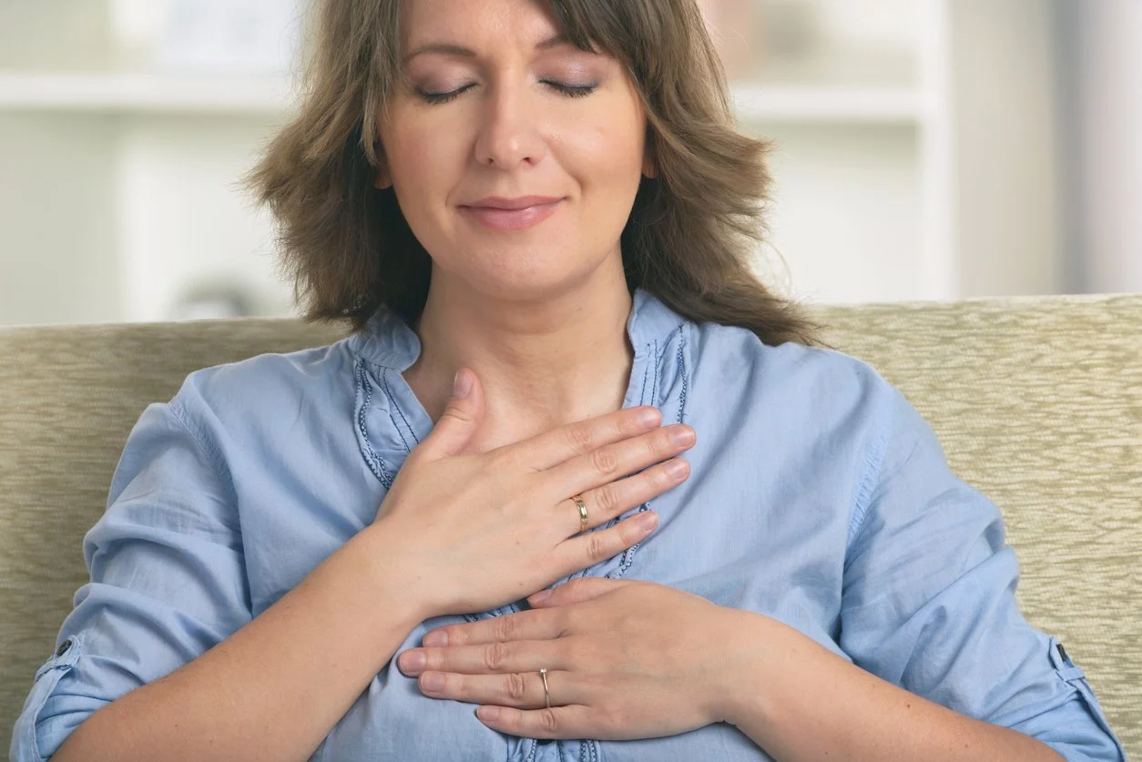 A woman with brown hair and a blue blouse is sitting on a beige couch with her eyes closed. She is holding her hands on her chest, appearing to be practicing deep breathing or meditation.