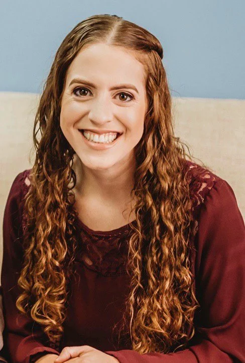 A young woman with long, curly, auburn hair, smiling, wearing a maroon top, sitting on a beige couch against a light blue wall.
