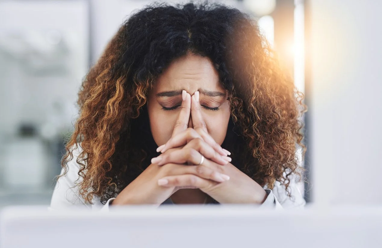 A shirtless man with tattoos, wearing a beaded necklace and bracelet, practicing meditation by a river in a natural setting with rocks and trees.