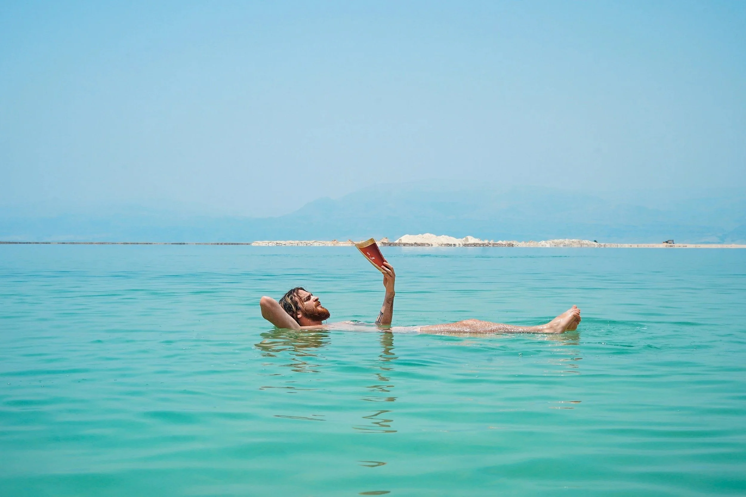 A man with a beard and long hair is floating in a body of turquoise water while reading a book under a clear blue sky.