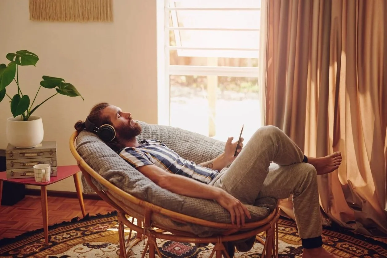 A man with a beard, wearing headphones, relaxing on a cushioned chair indoors, holding a smartphone, with sunlight coming through a window and beige curtains.