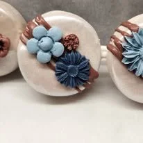 Three decorative round objects with blue and bronze floral decorations. The objects appear to be cookies or similar baked goods.