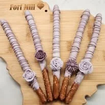 Five chocolate-dipped pretzel sticks decorated with purple and white beads and fabric flowers, arranged on a wooden cutting board.