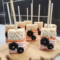 Five decorated marshmallow pops with pink stripes, black and pink flower embellishments, and white sprinkles on top, displayed on a wooden surface.