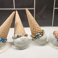 Four ice cream cones upside down on a surface, each coated with white frosting and decorated with colorful sprinkles.