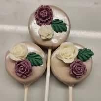 Lollipop pops decorated with icing flowers and leaves, resembling roses in lavender, white, and deep pink colors.
