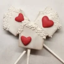 Decorated cookies shaped like hearts with red icing and white frosting, on white sticks.
