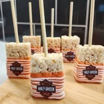 Six small cake pops with white frosting, orange drizzle, and a Harley-Davidson logo sticker, each on a wooden stick, arranged on a wooden surface.