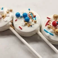 Three decorated cake pops with white icing on sticks, topped with colorful sprinkles and small candies.