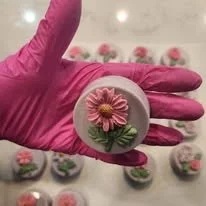 A gloved hand holding a round soap with a pink flower and green leaves embedded in it, with additional soaps featuring flower designs in the background.