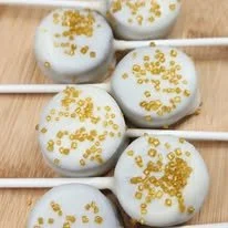 Five cake pops coated in white chocolate with gold sprinkles on a wooden surface.