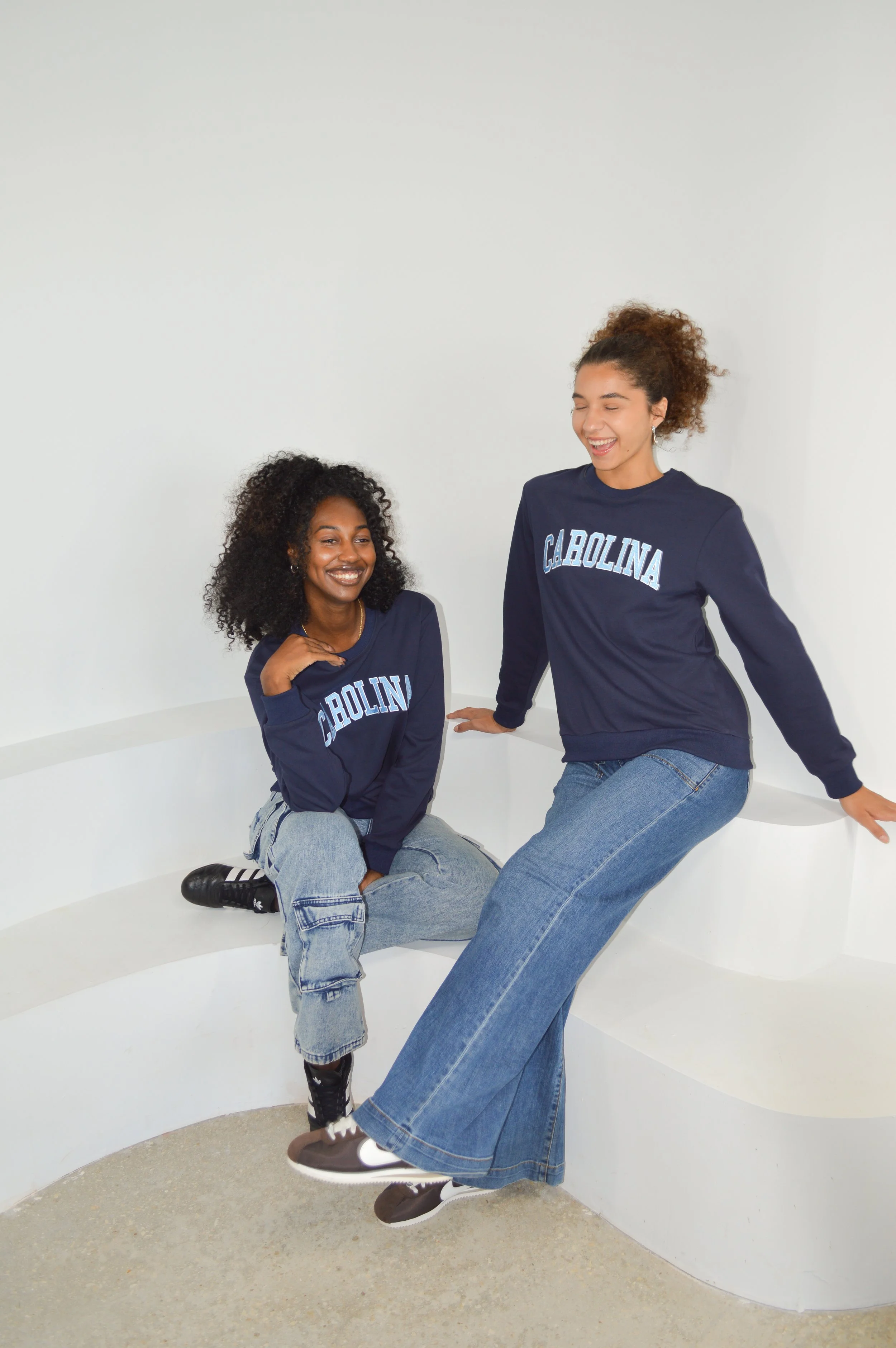 Two young women in navy blue sweatshirts with 'C. Carolina' written on them, sitting and laughing on a curved white bench in a minimalist white room.