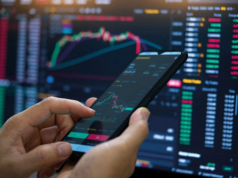 Person holding a smartphone displaying a stock trading app with a financial chart, in front of a large screen showing stock market data and graphs.