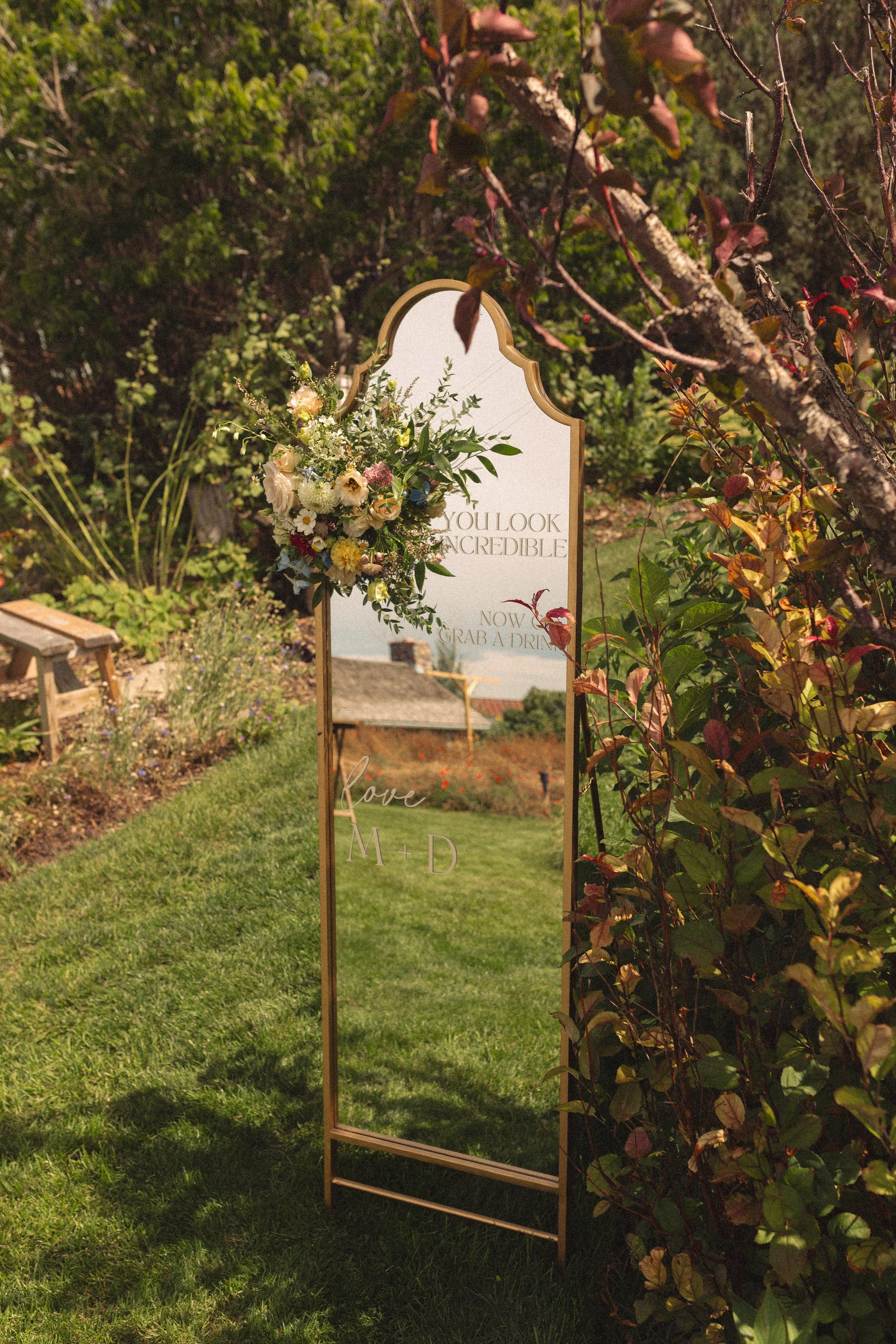 A tall, narrow mirror with a gold frame placed outdoors on a grassy area, decorated with a floral arrangement on its upper left corner, reading 'You look incredible, now grab a drink.' In the background, there are trees, bushes, and a bench, suggesti