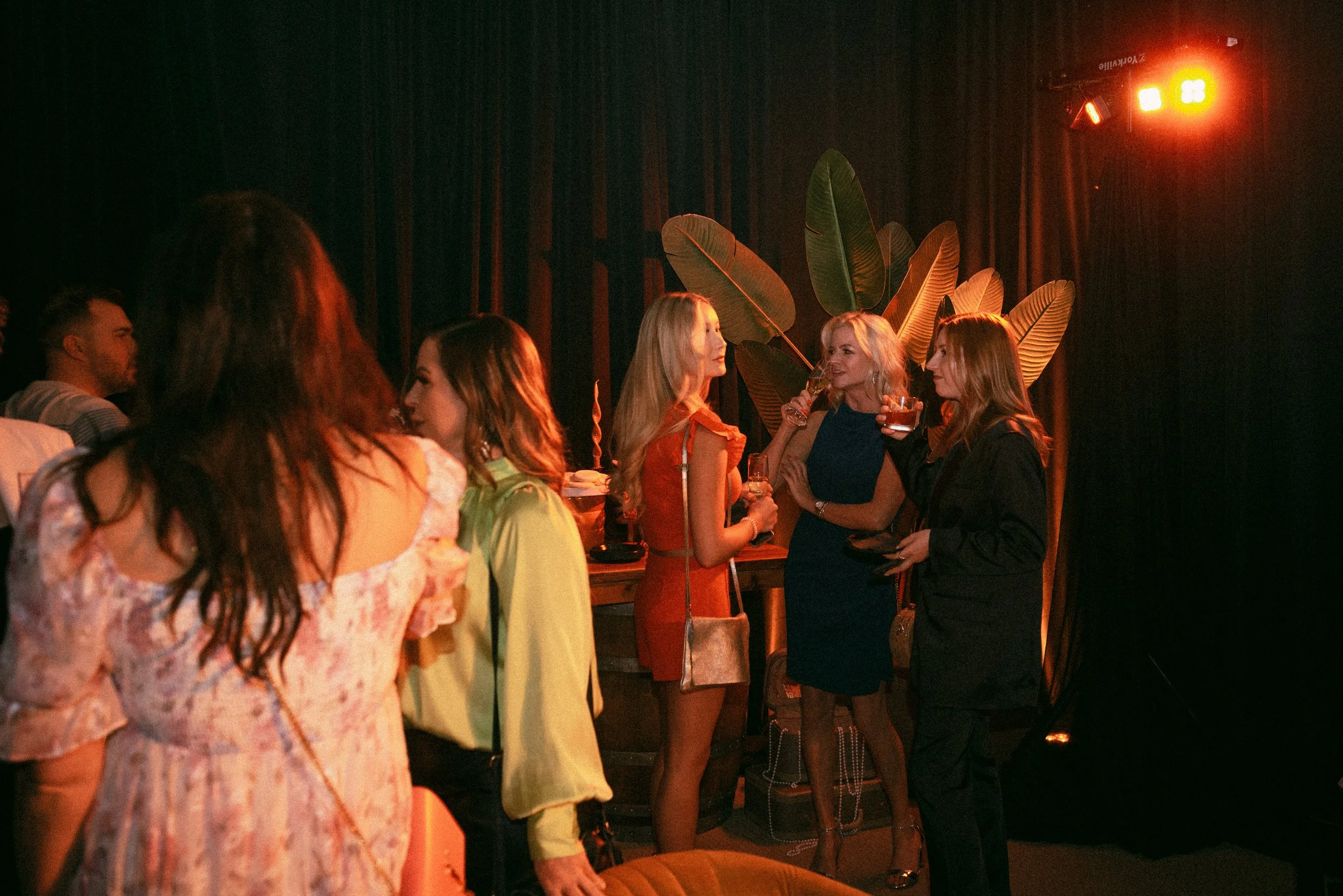 Group of women in cocktail dresses standing and chatting at a party with dim lighting and large decorative plants in the background.