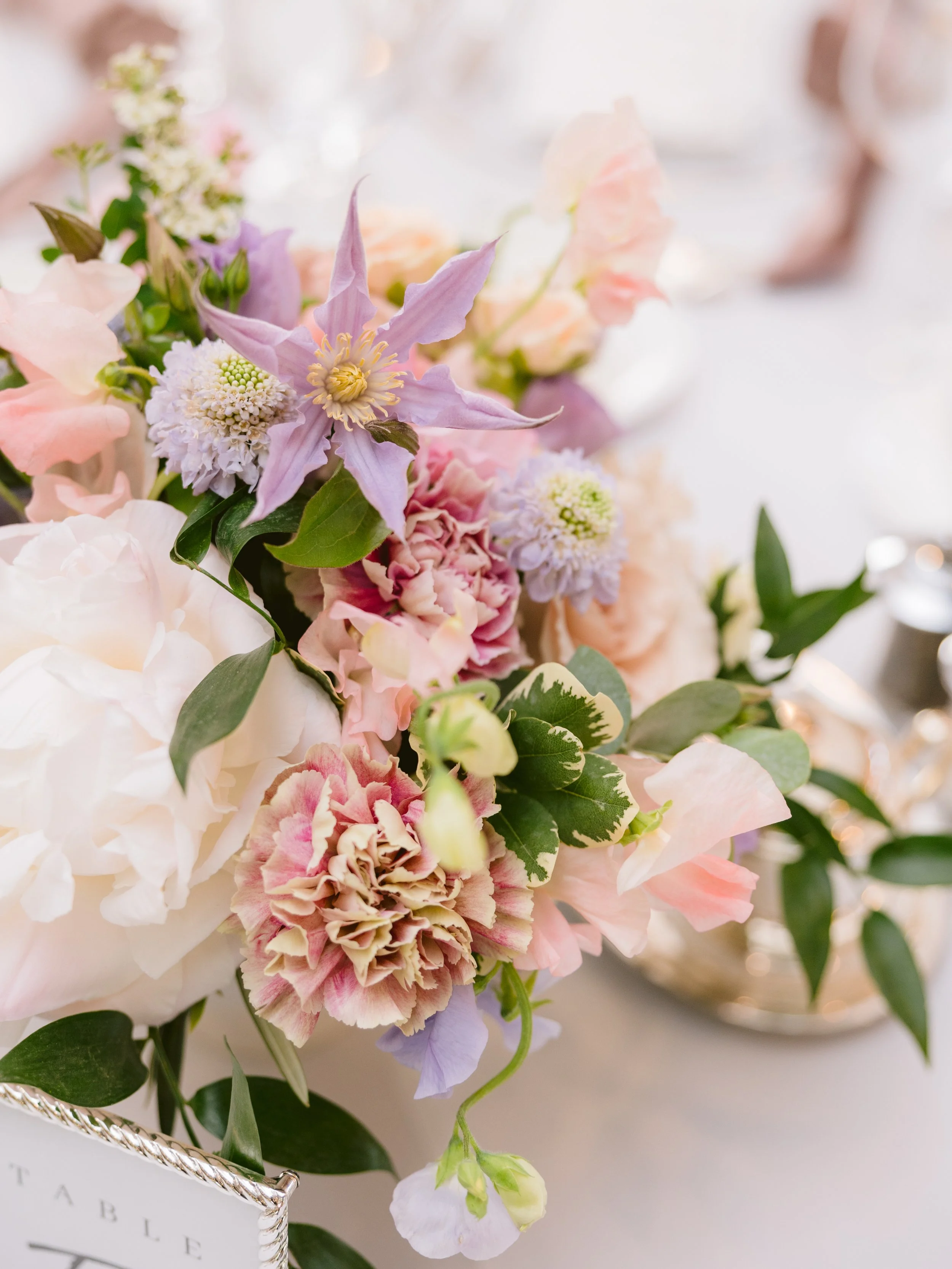 A close-up of a pastel-colored flower arrangement with pink, purple, white, and green flowers and leaves, placed on a table with a silver-gilded table setting in the background.