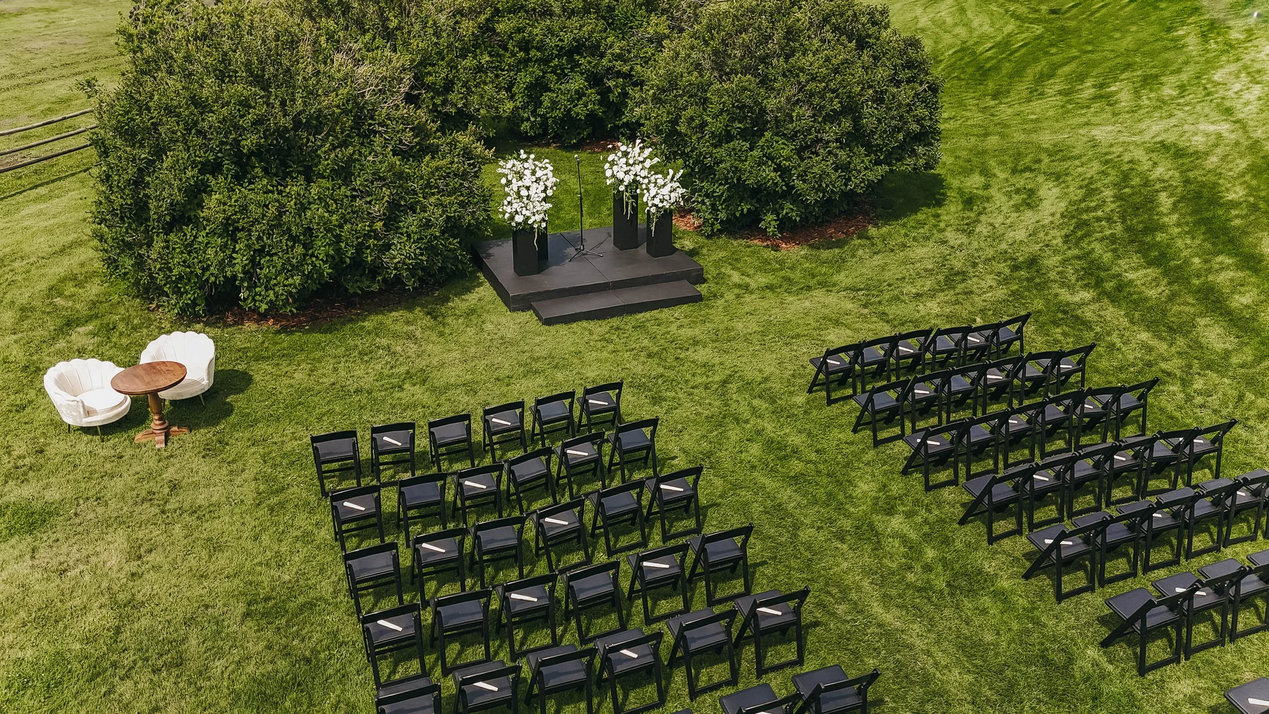 An outdoor wedding ceremony setup on a green lawn with black chairs arranged in rows facing a small stage with a microphone and two black pedestals holding white flower arrangements. There is a small seating area with two white chairs and a round woo