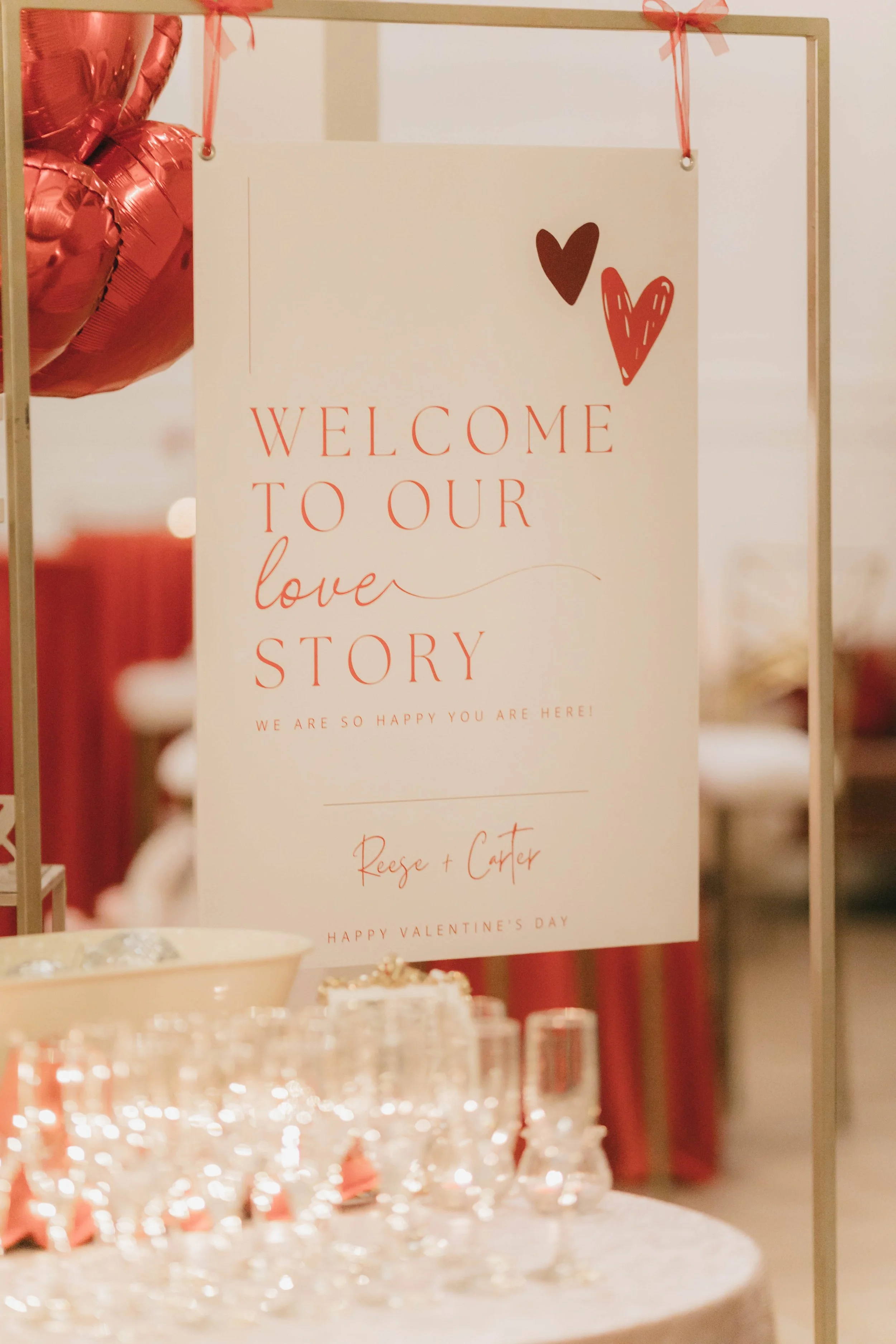 Valentine's Day event sign that reads 'Welcome to our love story, Reese + Carter, Happy Valentine's Day,' decorated with red and brown hearts, with glasses and bowls of snacks in the foreground.