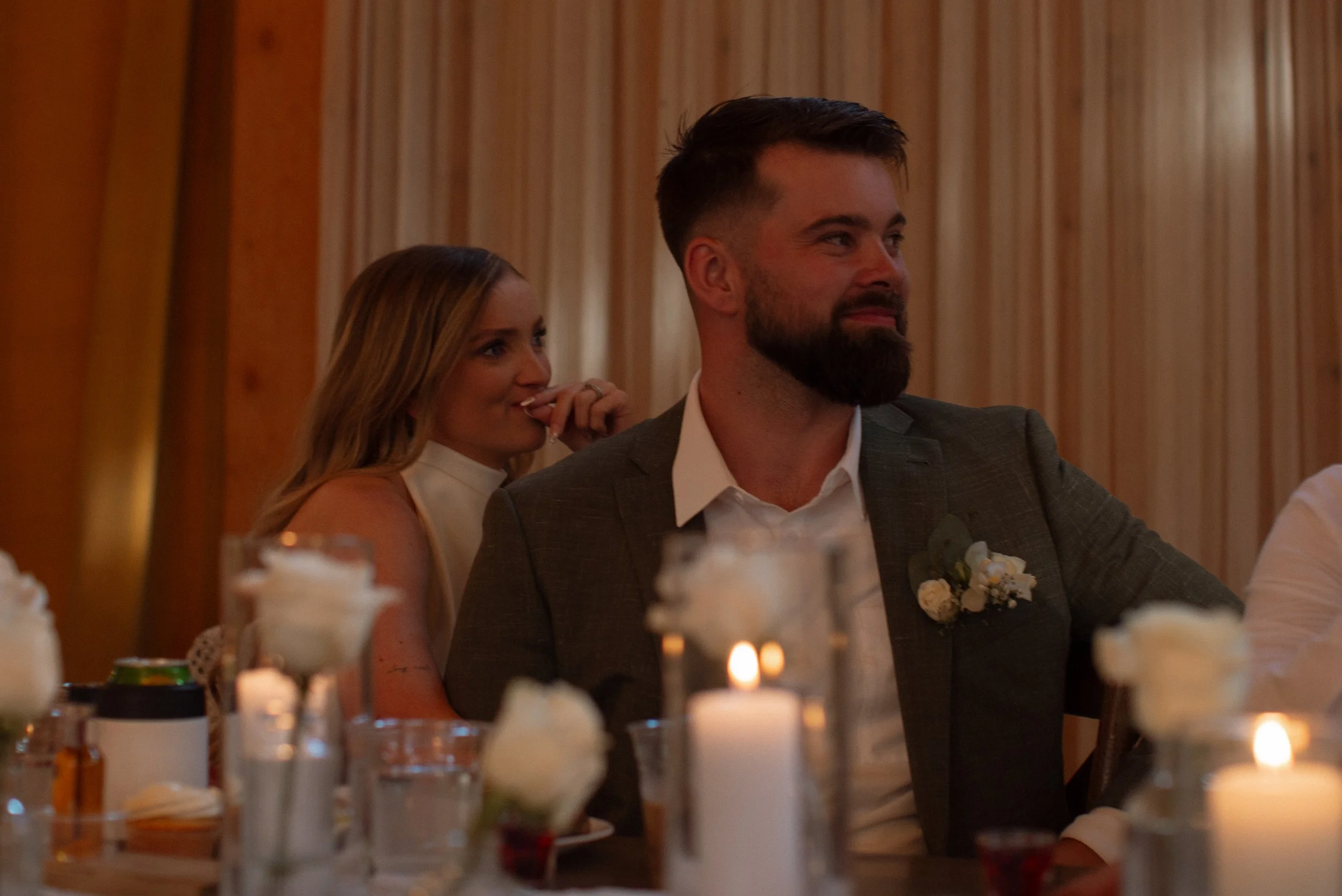A man in a suit with a boutonniere sits at a table with candles, joined by a woman in a white dress, at a formal event or wedding reception.