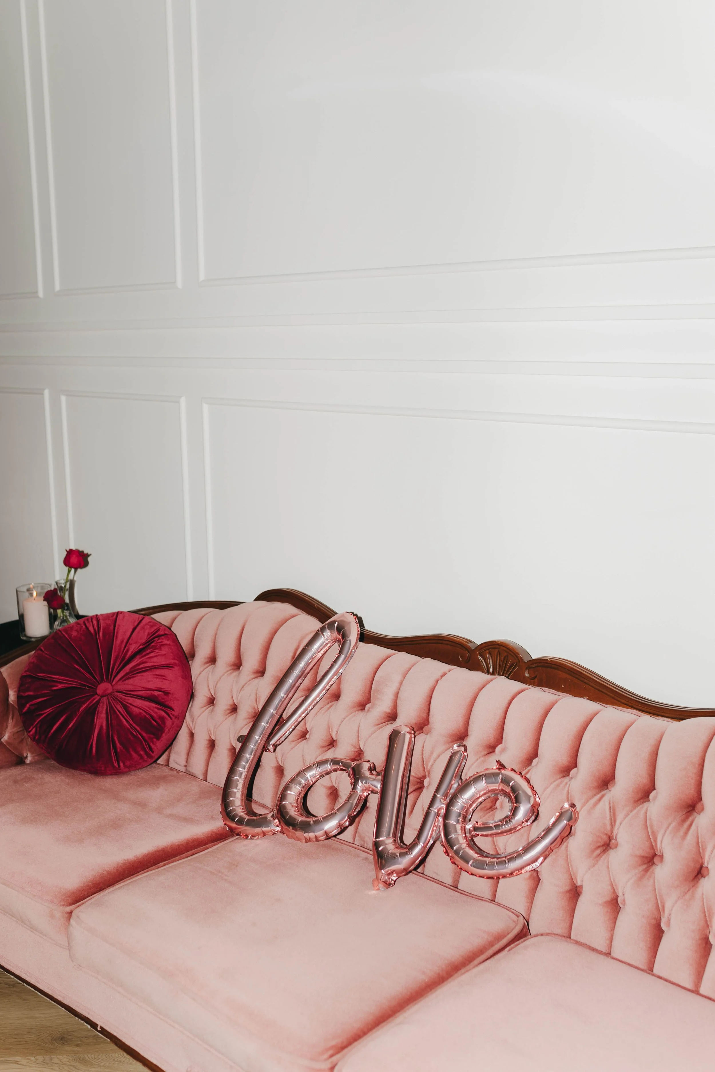 A pink tufted vintage sofa with a red round pillow, a small table with a candle and red roses, and decorative metallic balloon letters spelling 'Love' resting on the sofa. White paneled wall background.