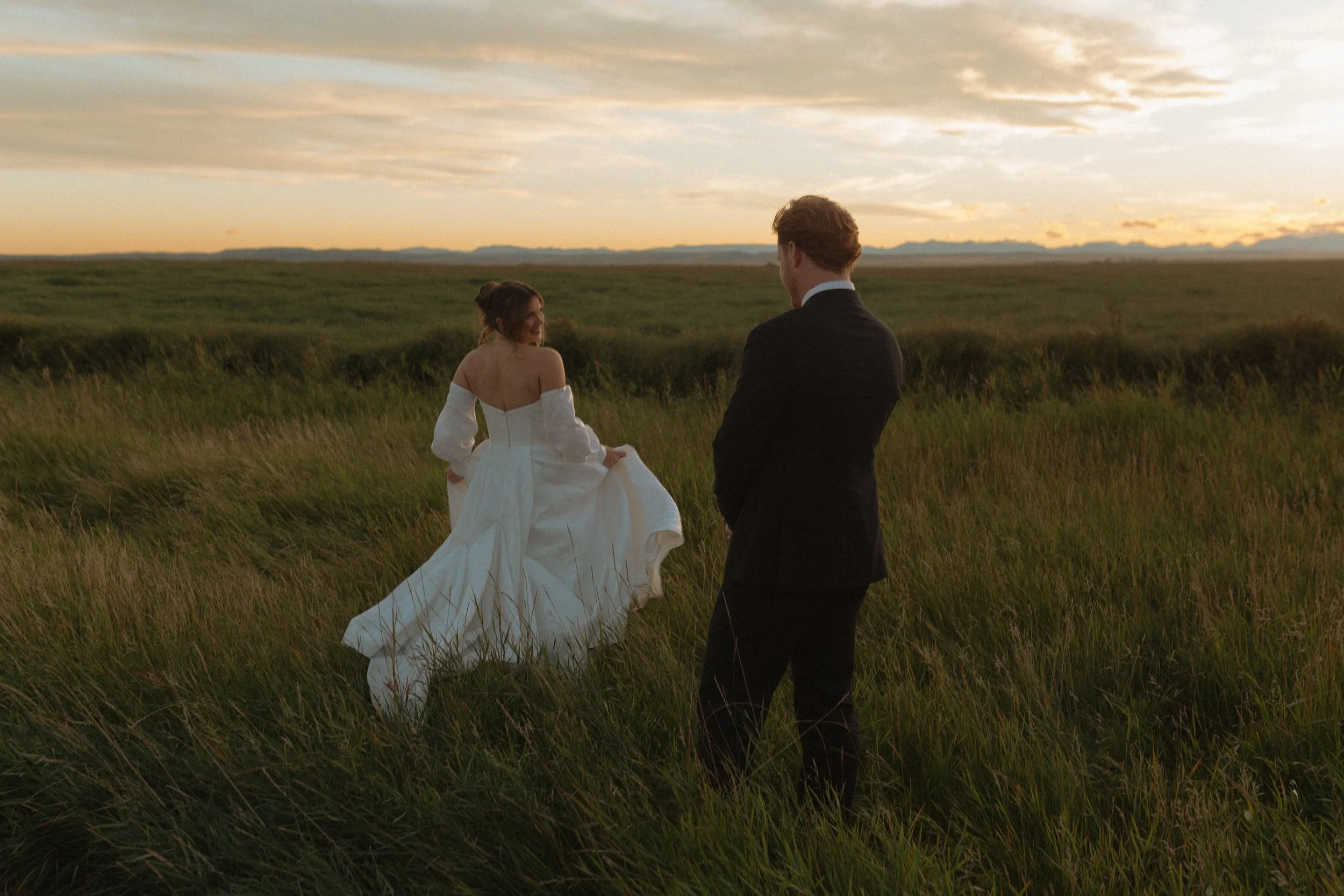 A woman in a wedding dress and a man in a black suit standing in a grassy field at sunset, looking at each other.