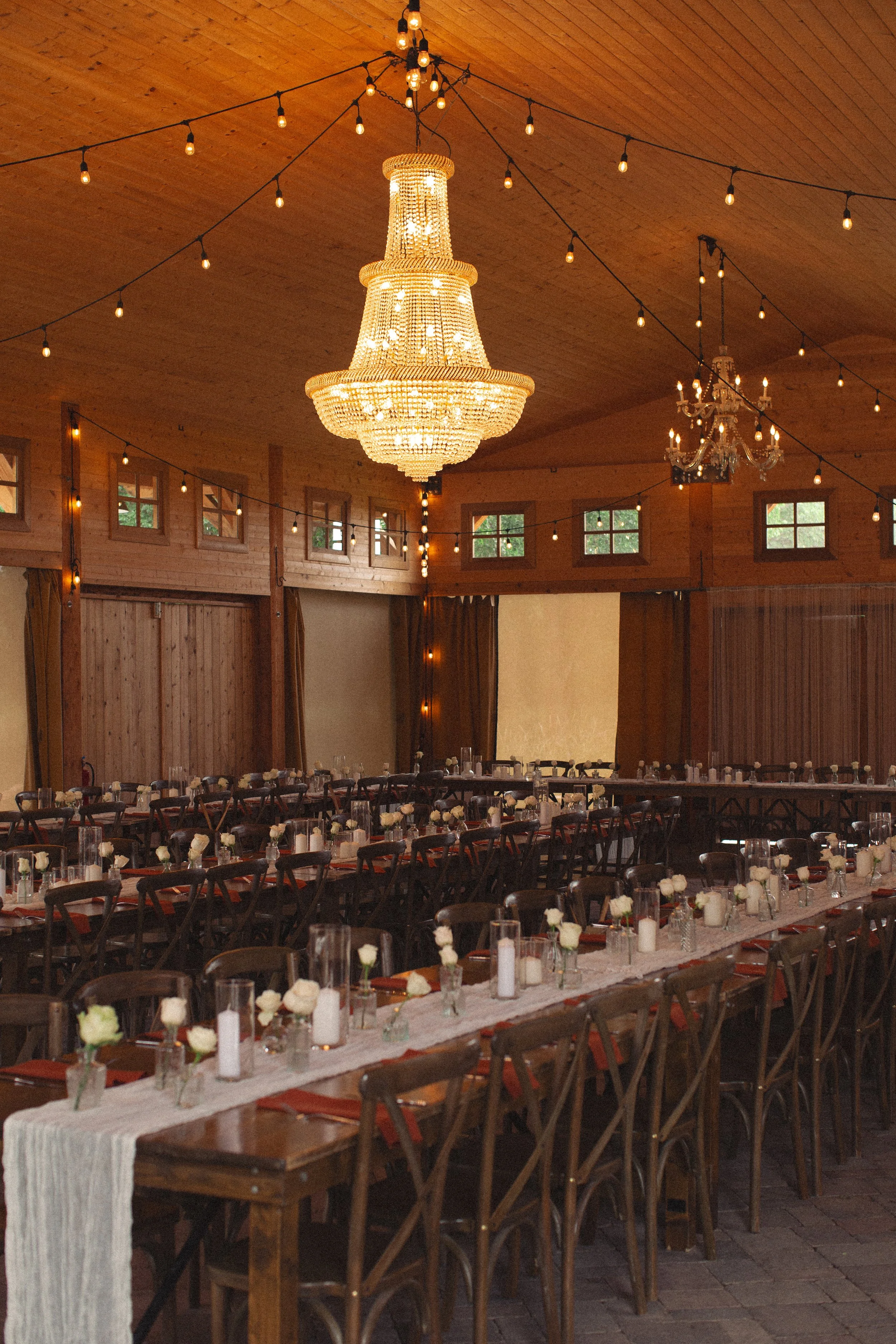 Elegant rustic wedding reception hall with long tables decorated with white flowers and candles, wood-paneled walls, large chandeliers, and string lights hanging from the ceiling.