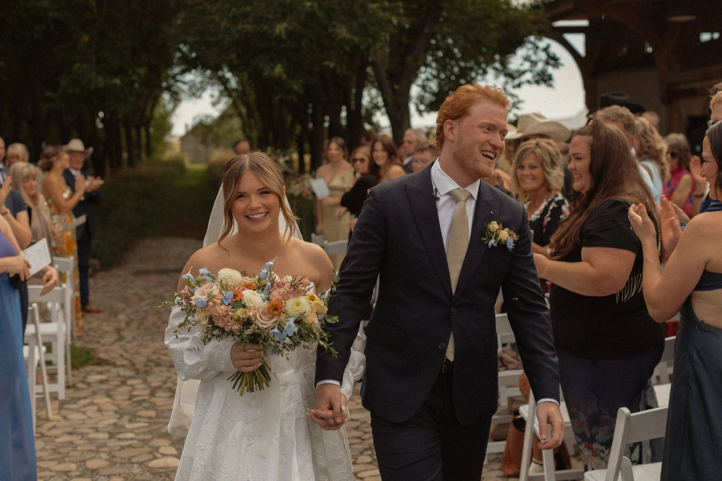 A bride and groom walking hand in hand, smiling after their wedding ceremony outdoors, surrounded by friends and family.