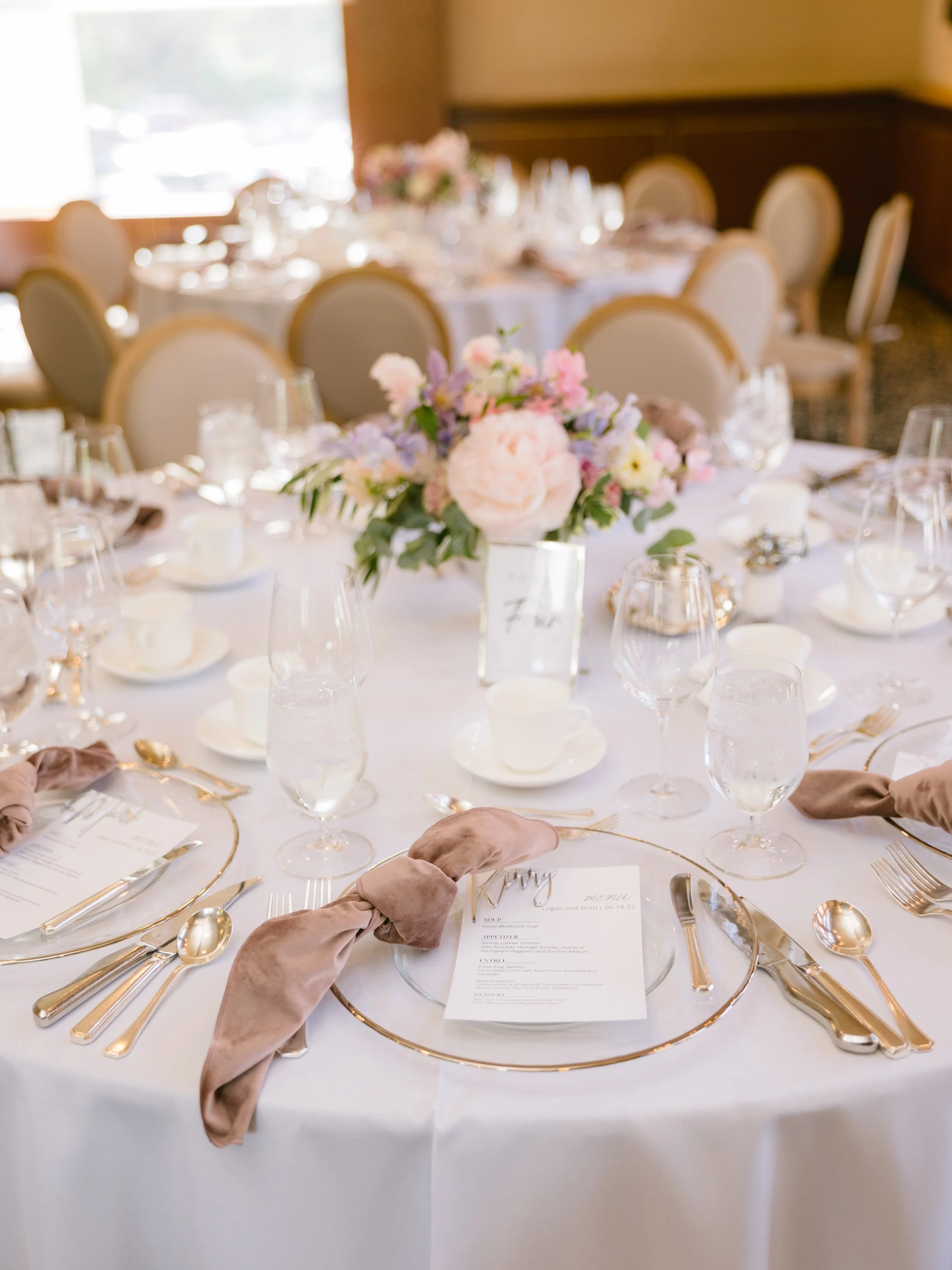 Elegant wedding reception table with a floral centerpiece, gold cutlery, place cards, menus, and water glasses, set for dining.