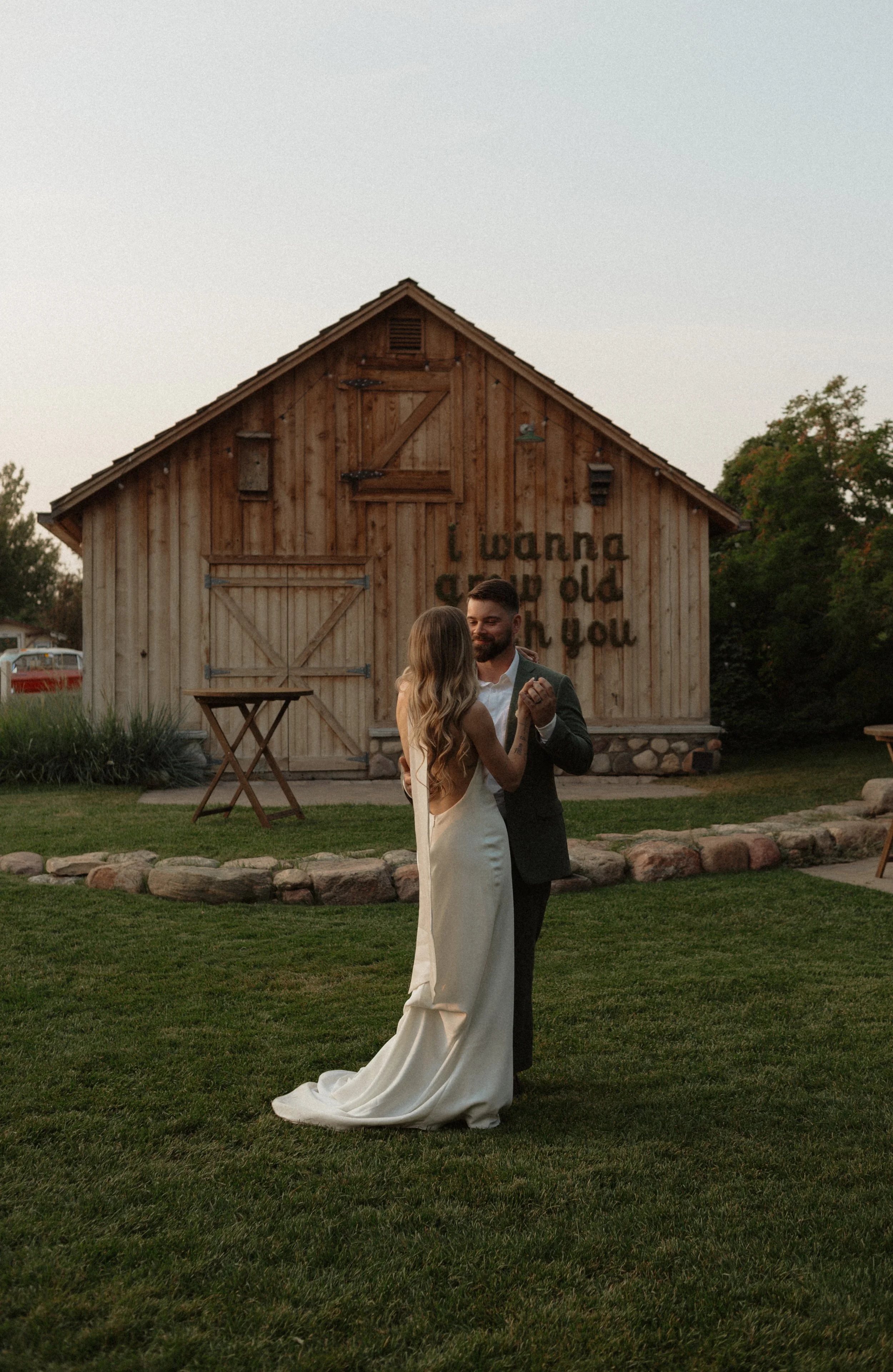 A couple dancing outside at sunset, in wedding attire, with a rustic wooden barn in the background, partially covered by trees.